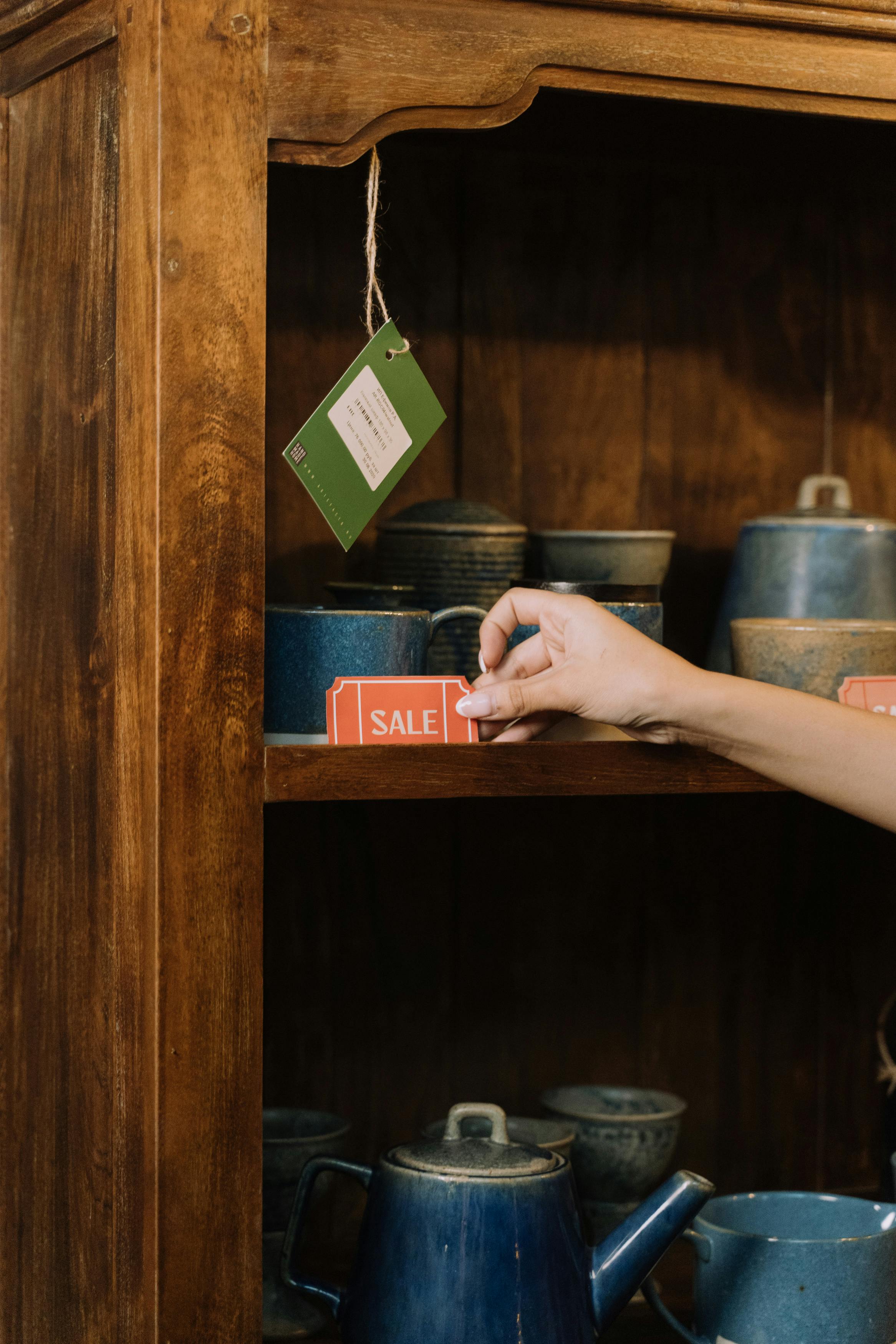 Hand adjusting sale tag on a shelf with blue pottery in a rustic wooden setting.