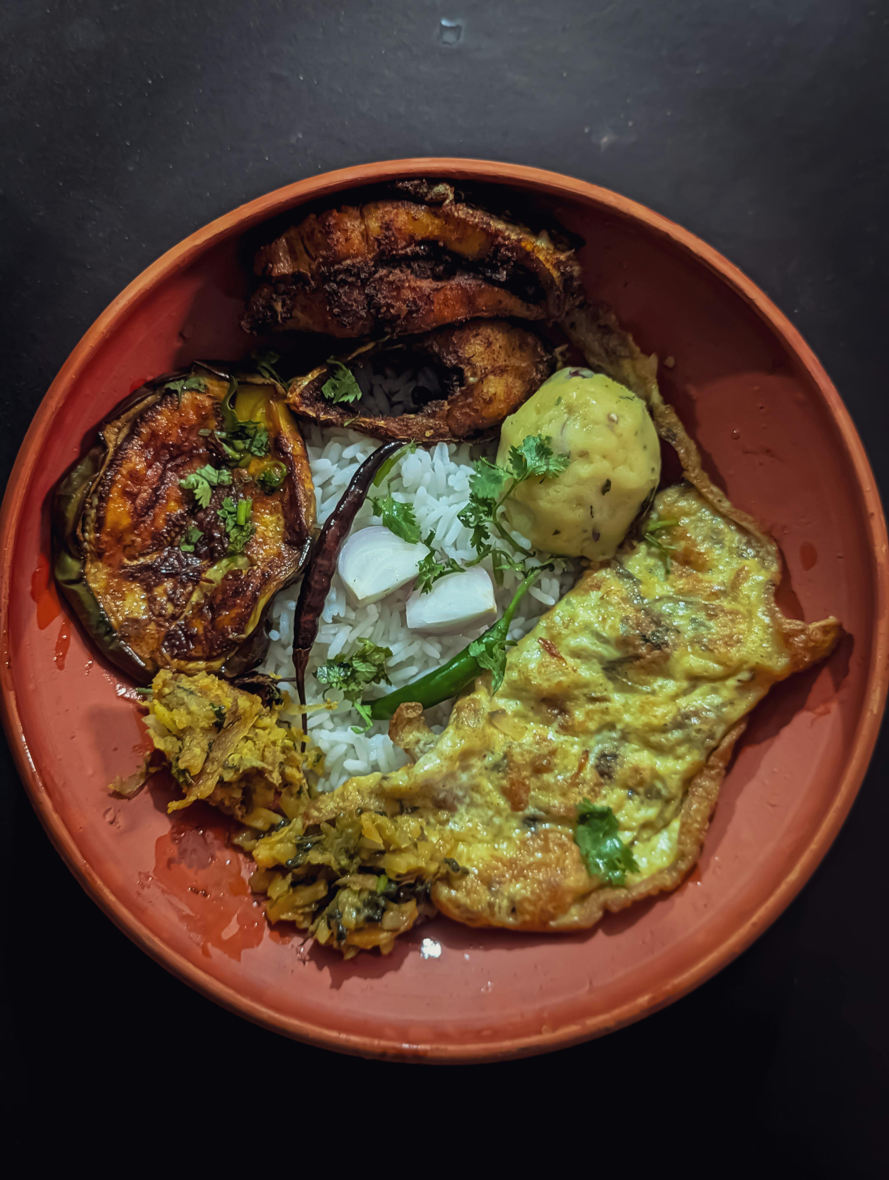 Delicious Bangladeshi meal with rice, fried fish, omelet, and vegetables served in a clay dish.
