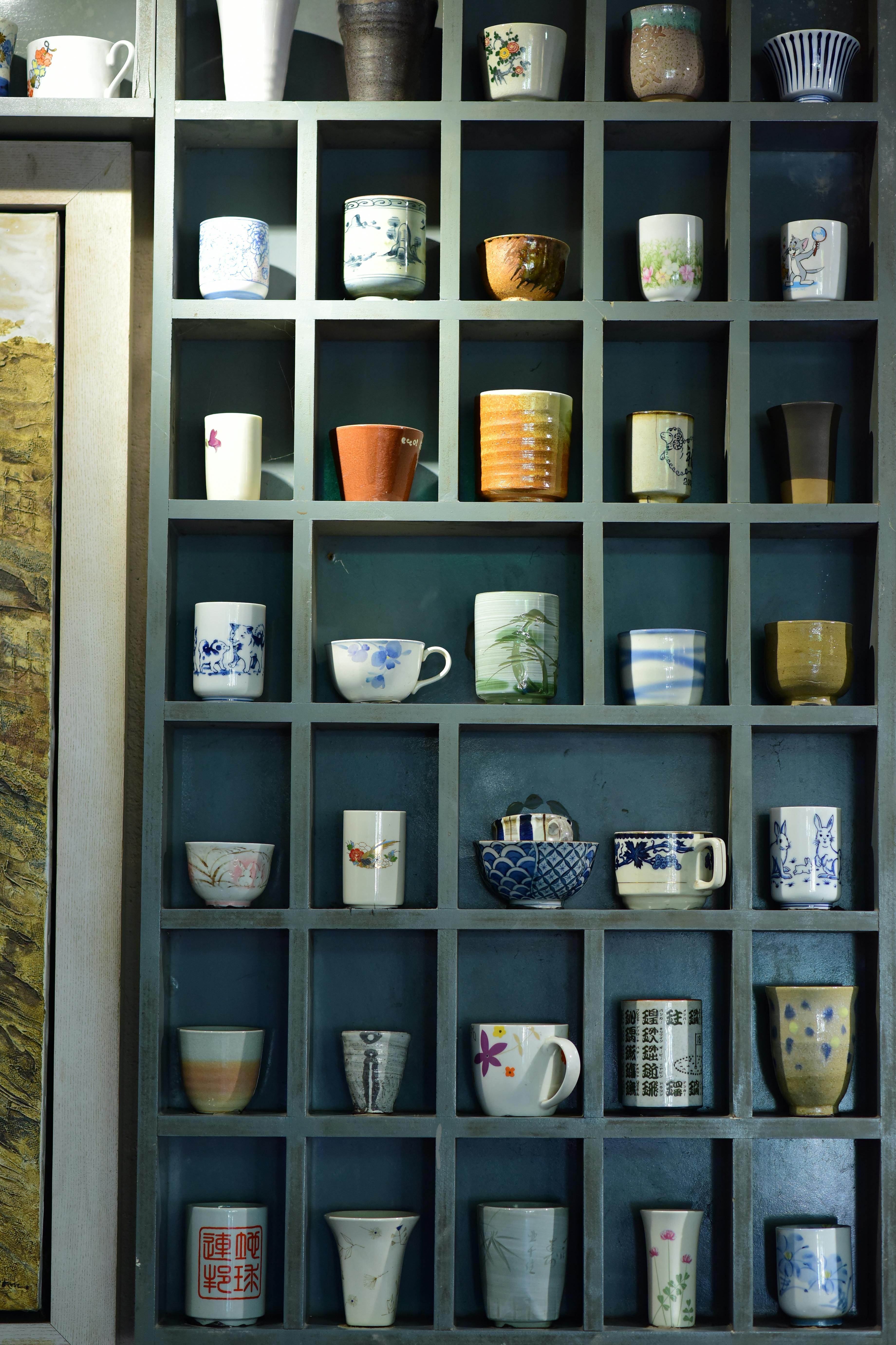 A variety of decorative ceramic cups and mugs arranged on a display shelf, showcasing diverse designs and colors.