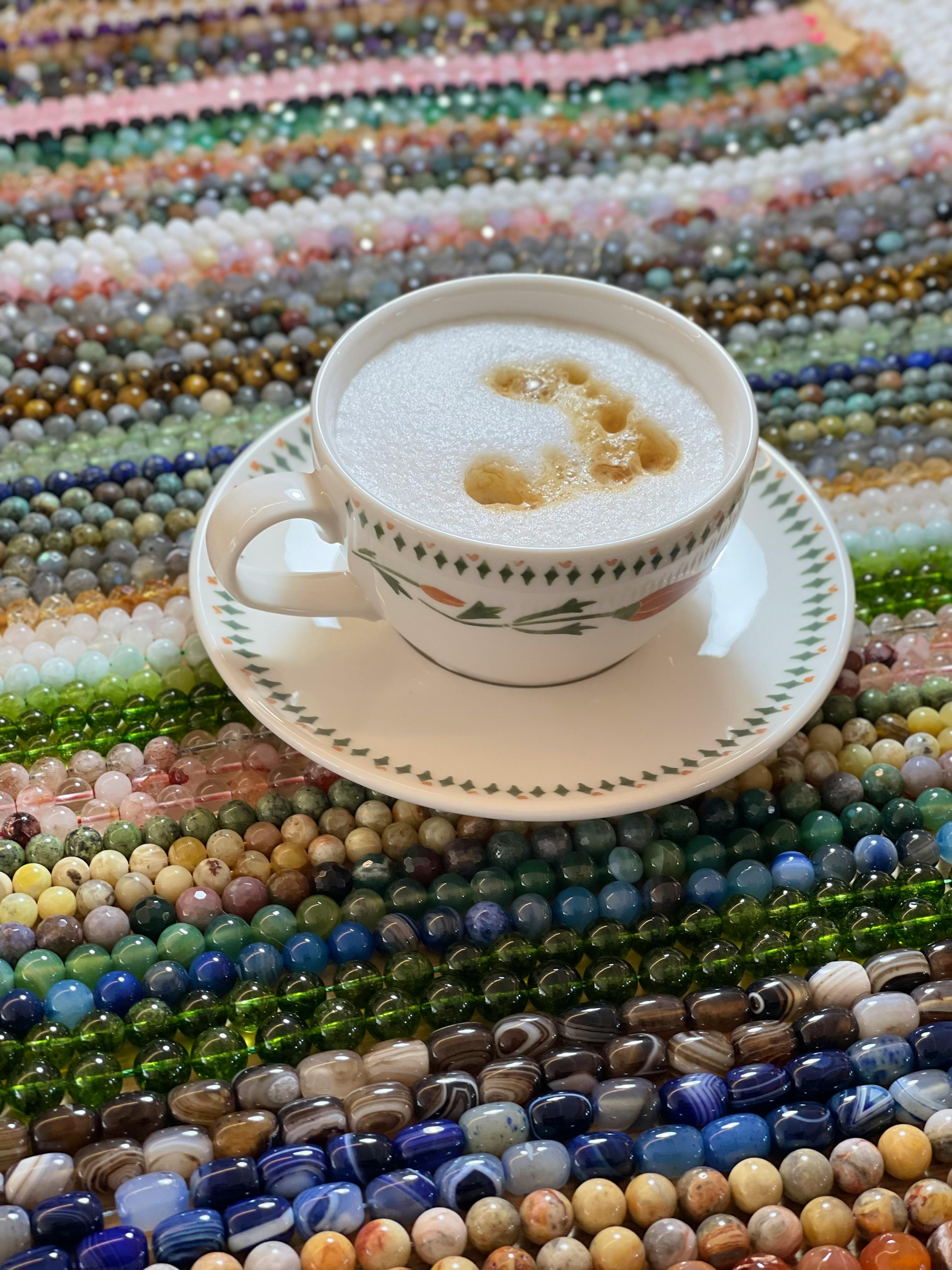 A frothy latte in a floral porcelain cup sits on a vibrant bead surface.