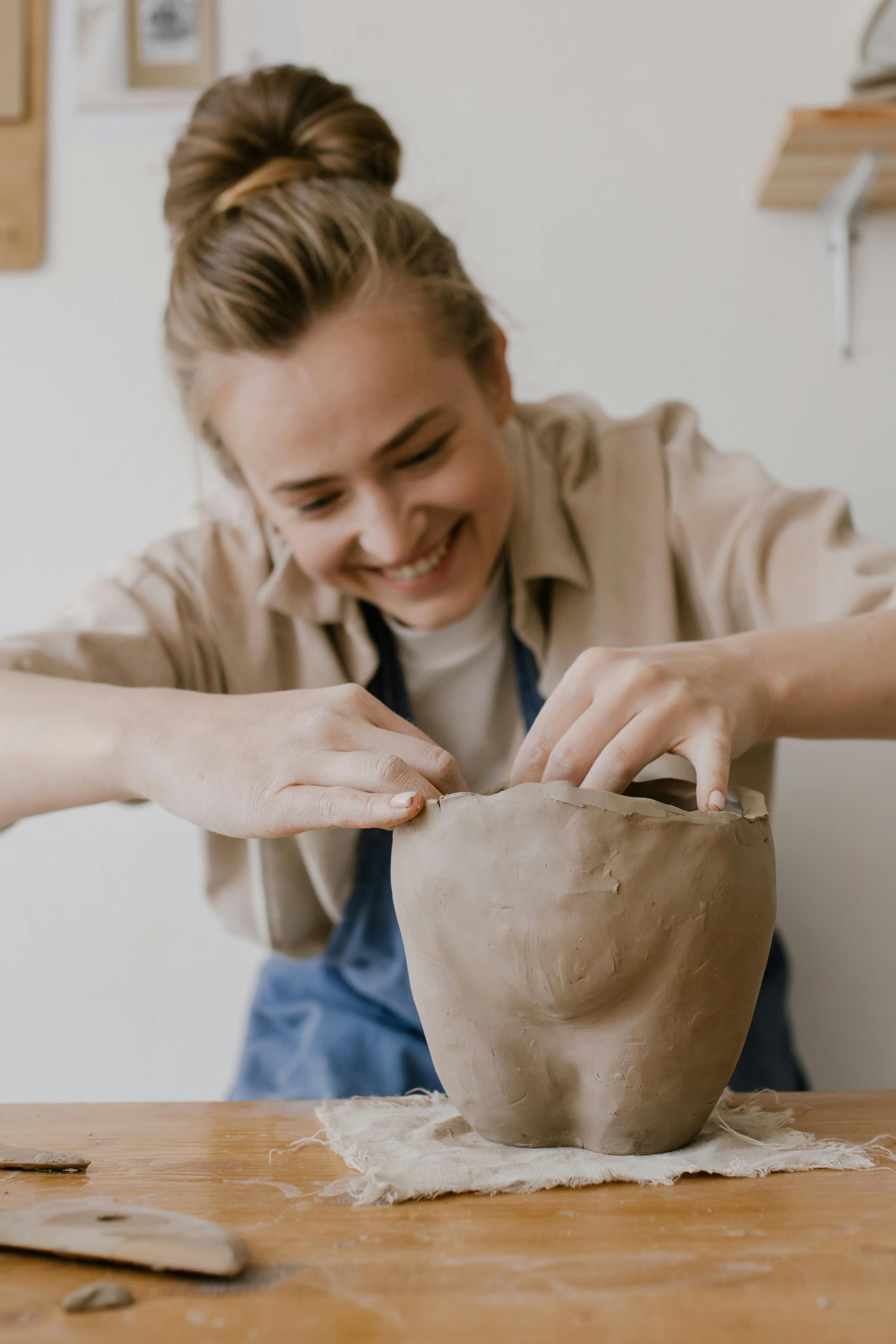 Cheerful woman sculptor shaping clay pot at indoor workshop table.