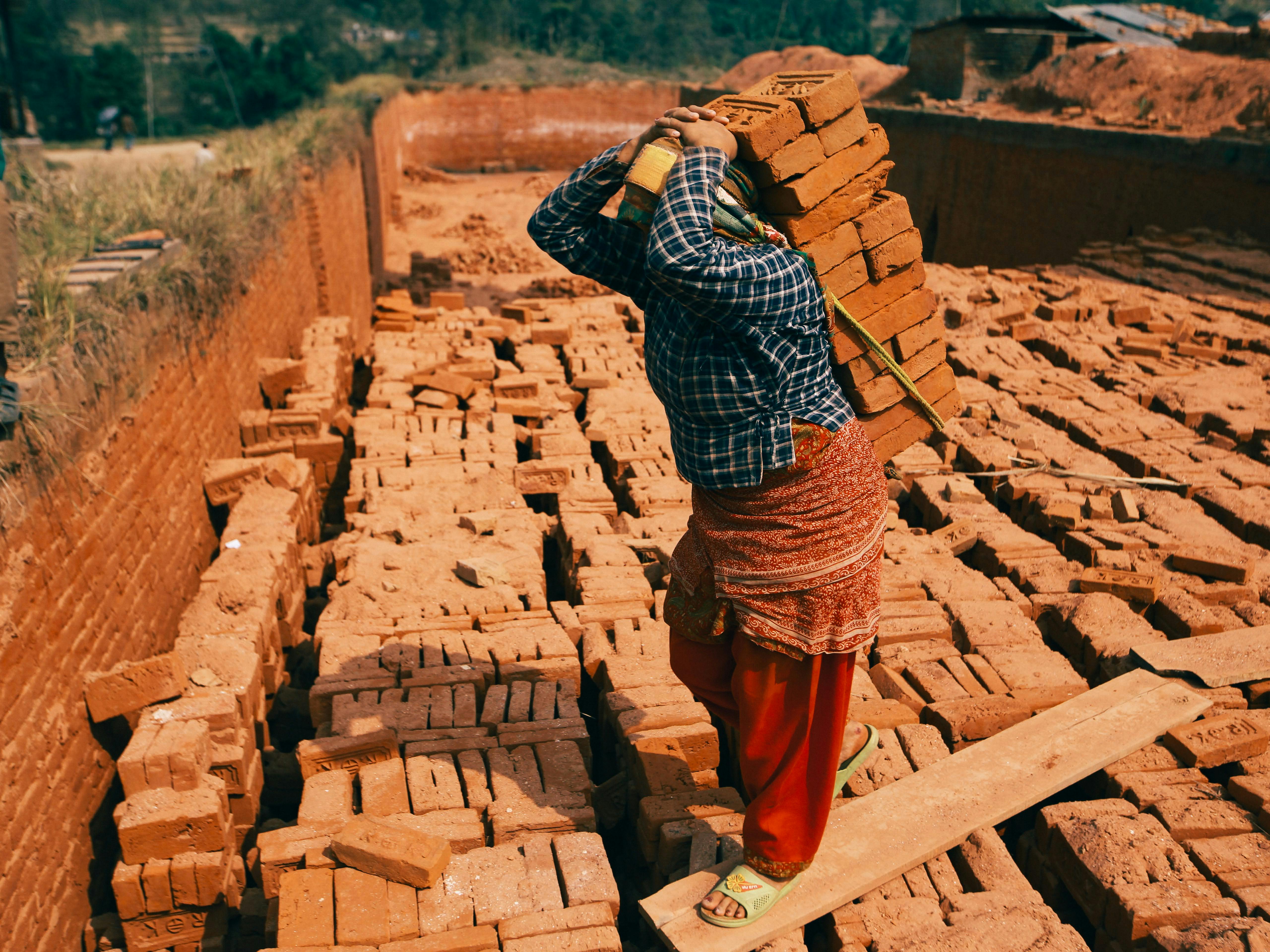 A woman works in a brick construction site, carrying bricks across a plank in a sunny outdoor setting.