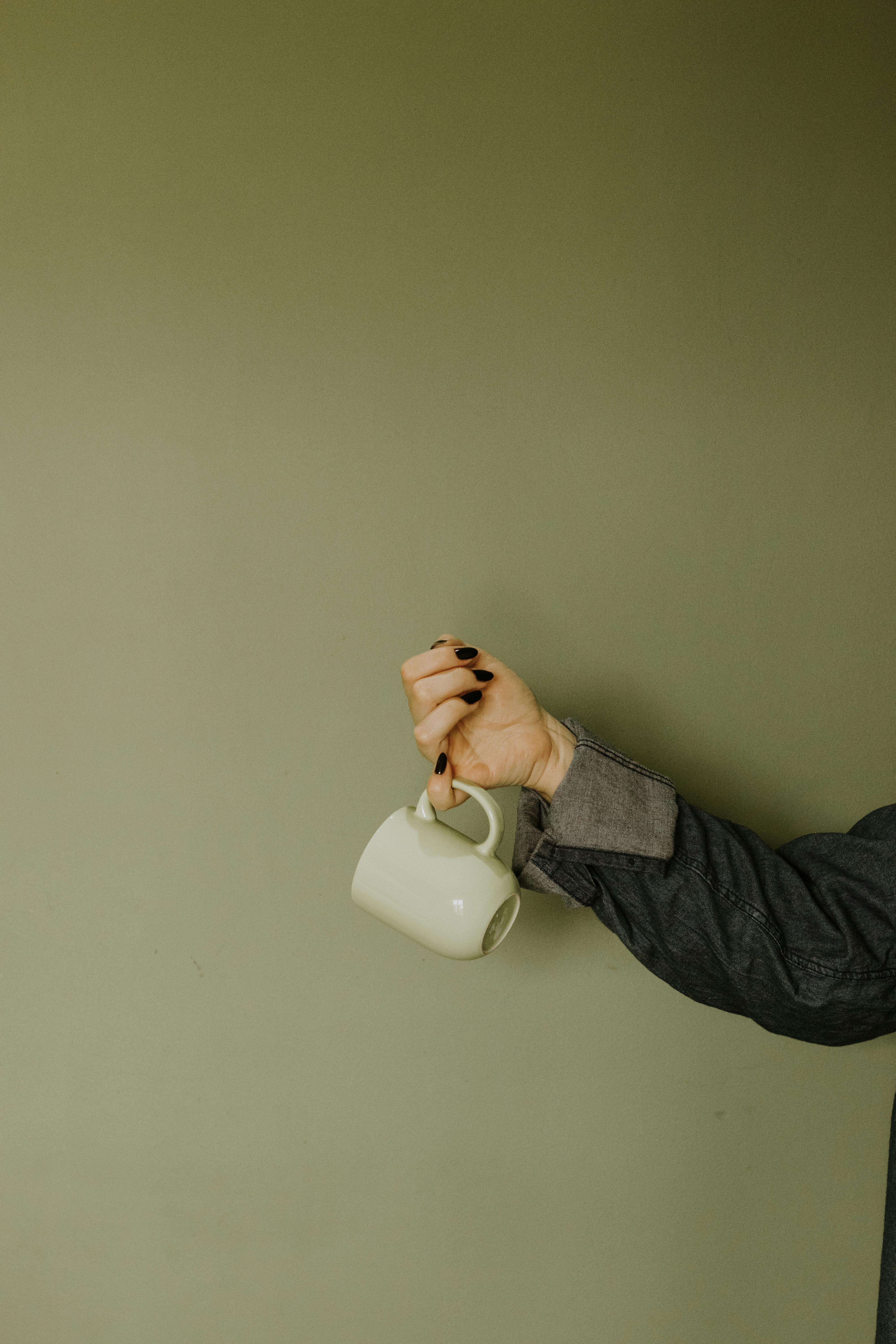 A stylish ceramic mug held by a woman's hand with dark nail polish in front of a muted olive green background.