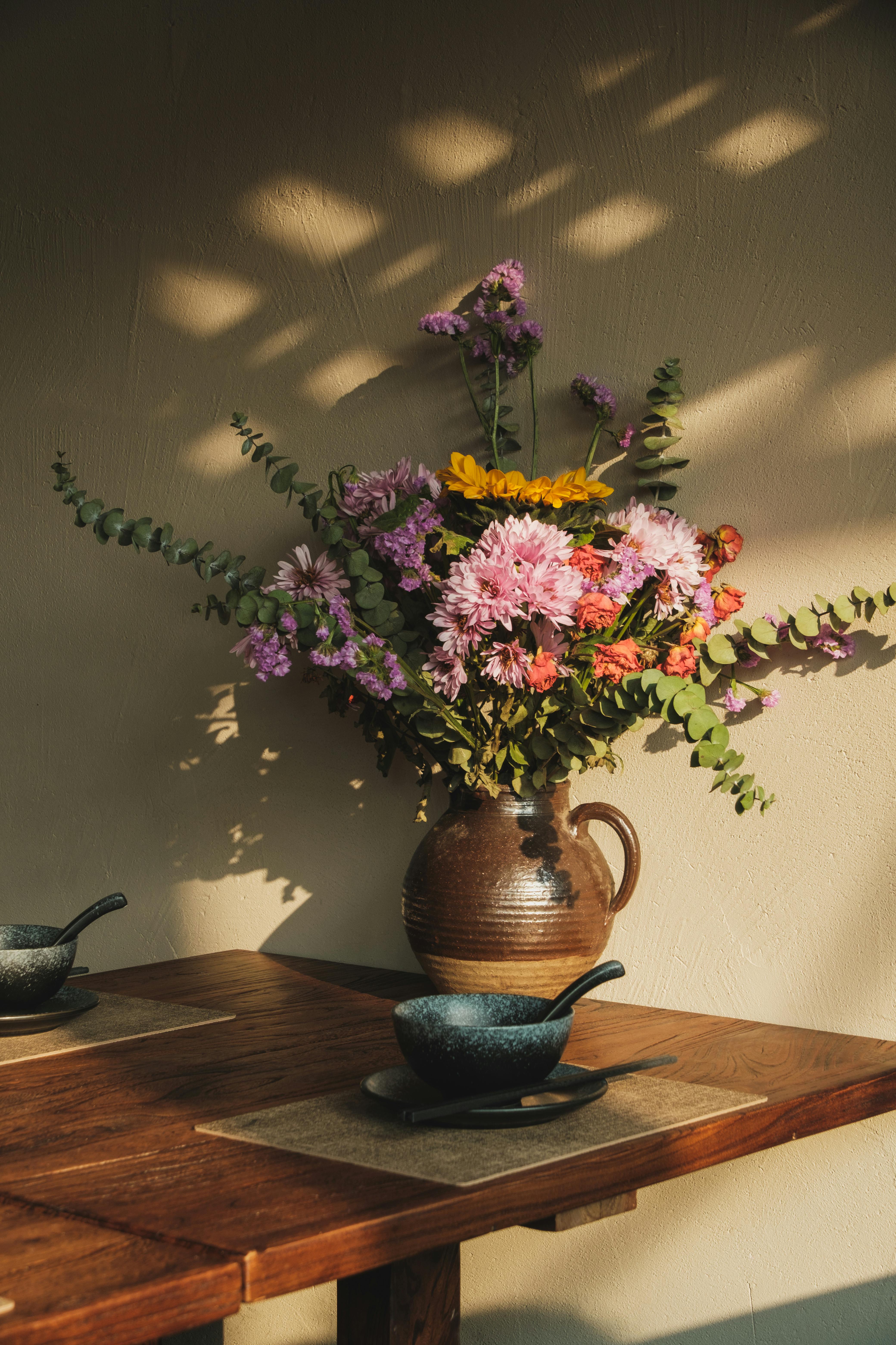 A rustic ceramic vase with vibrant flowers on a sunlit wooden table.