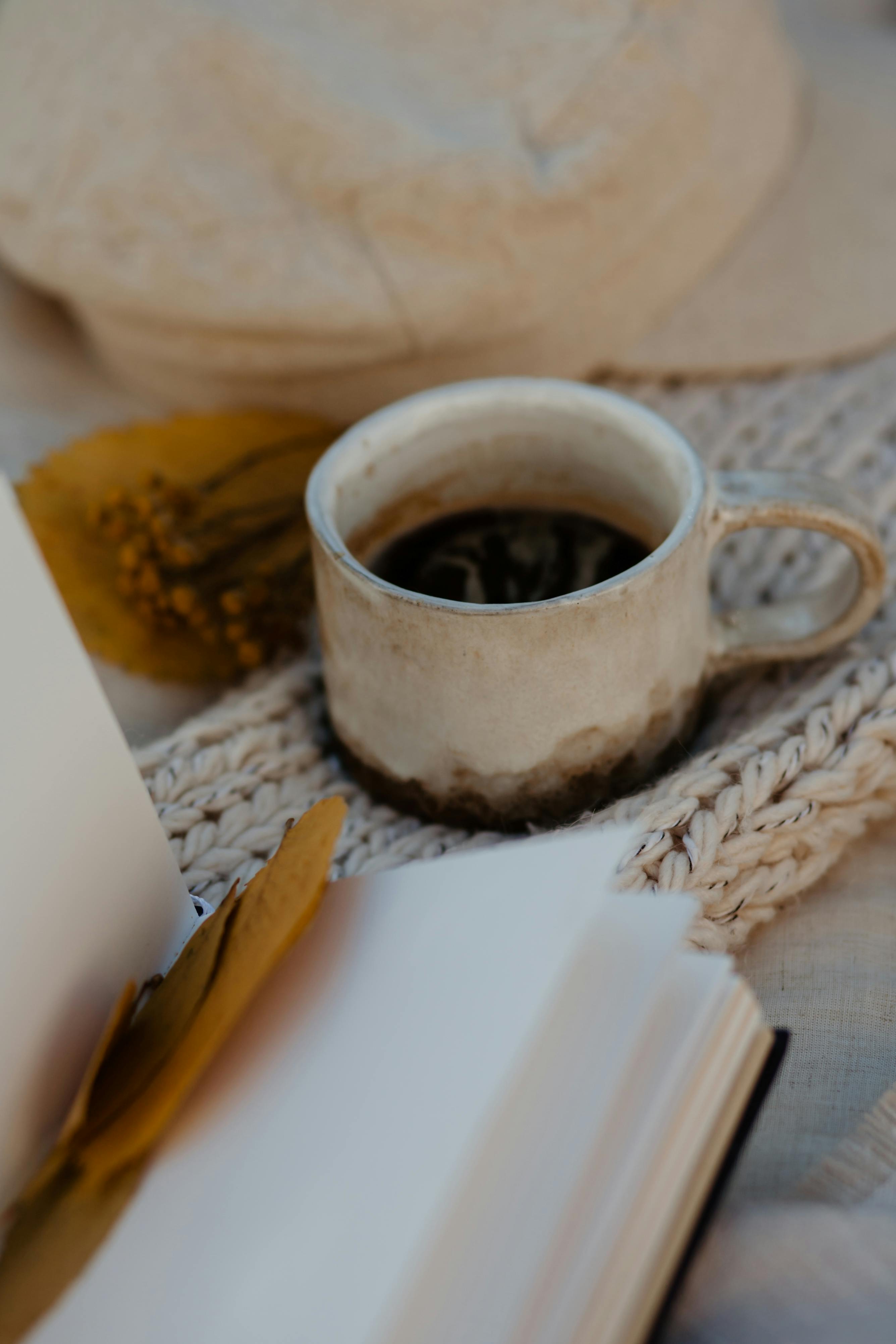 A rustic ceramic coffee cup sits beside an open book, creating a cozy still life scene.