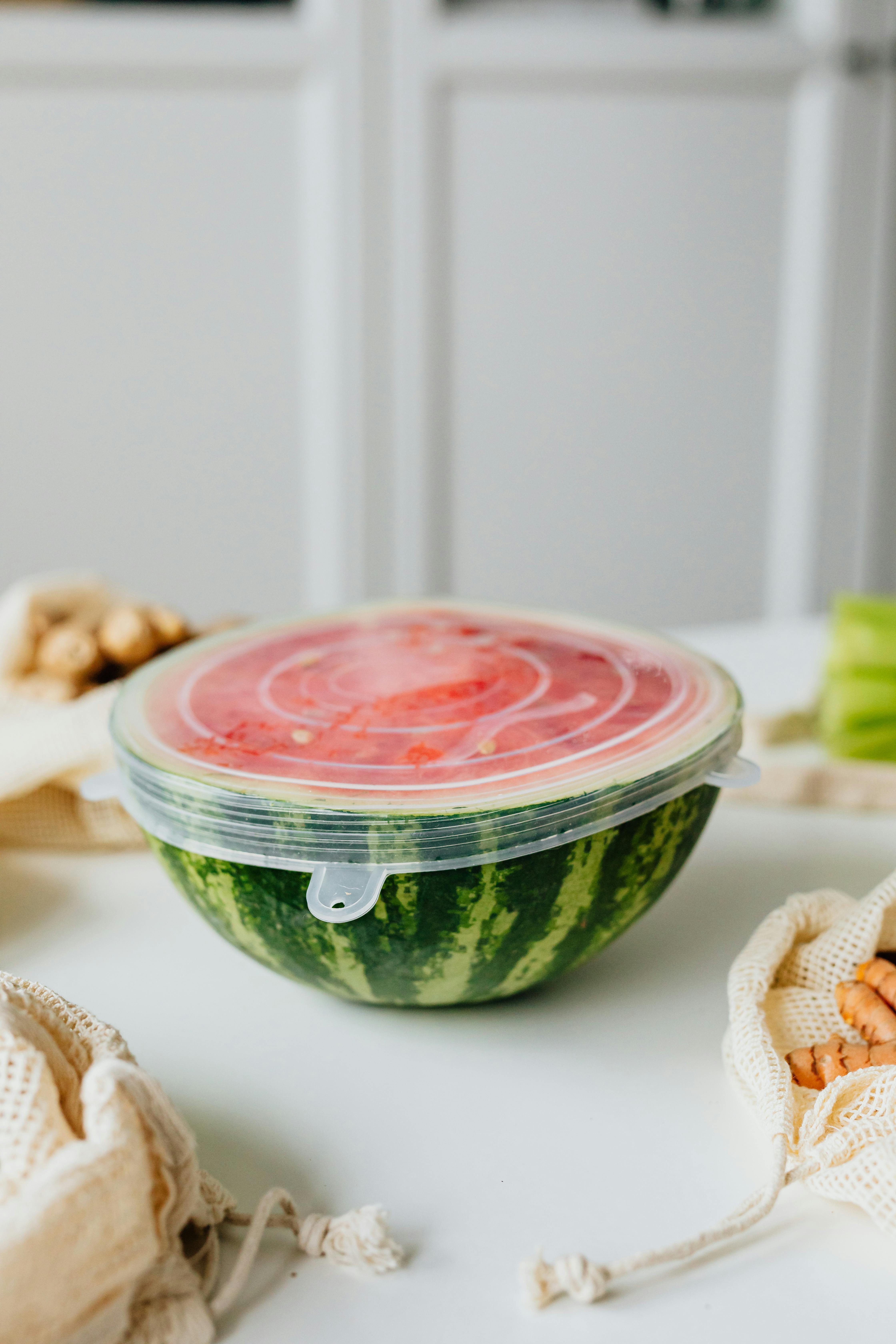 Sliced watermelon in airtight cover on kitchen table with reusable bags and snacks.