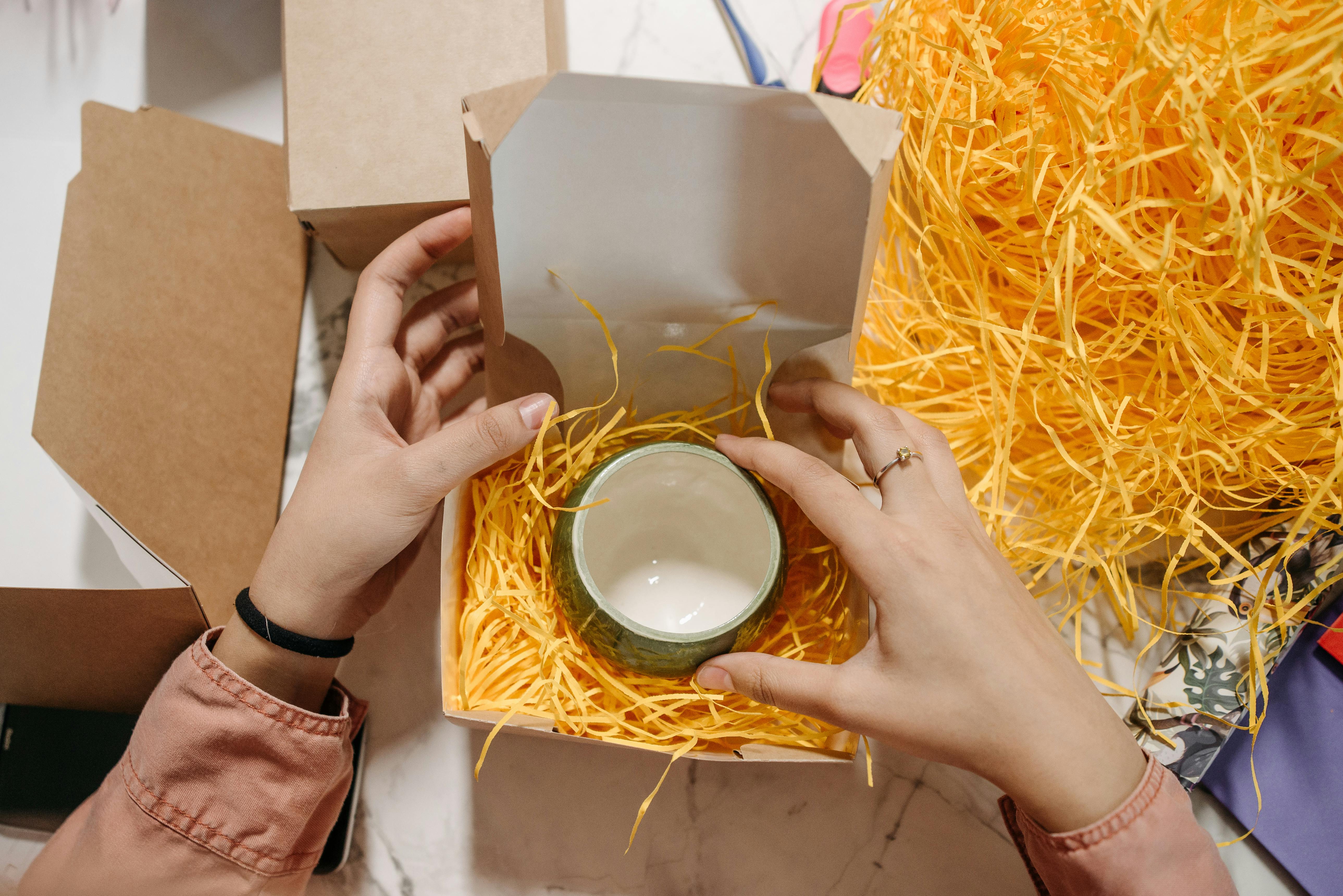 Close-up of hands unboxing a ceramic item with shredded packing material.