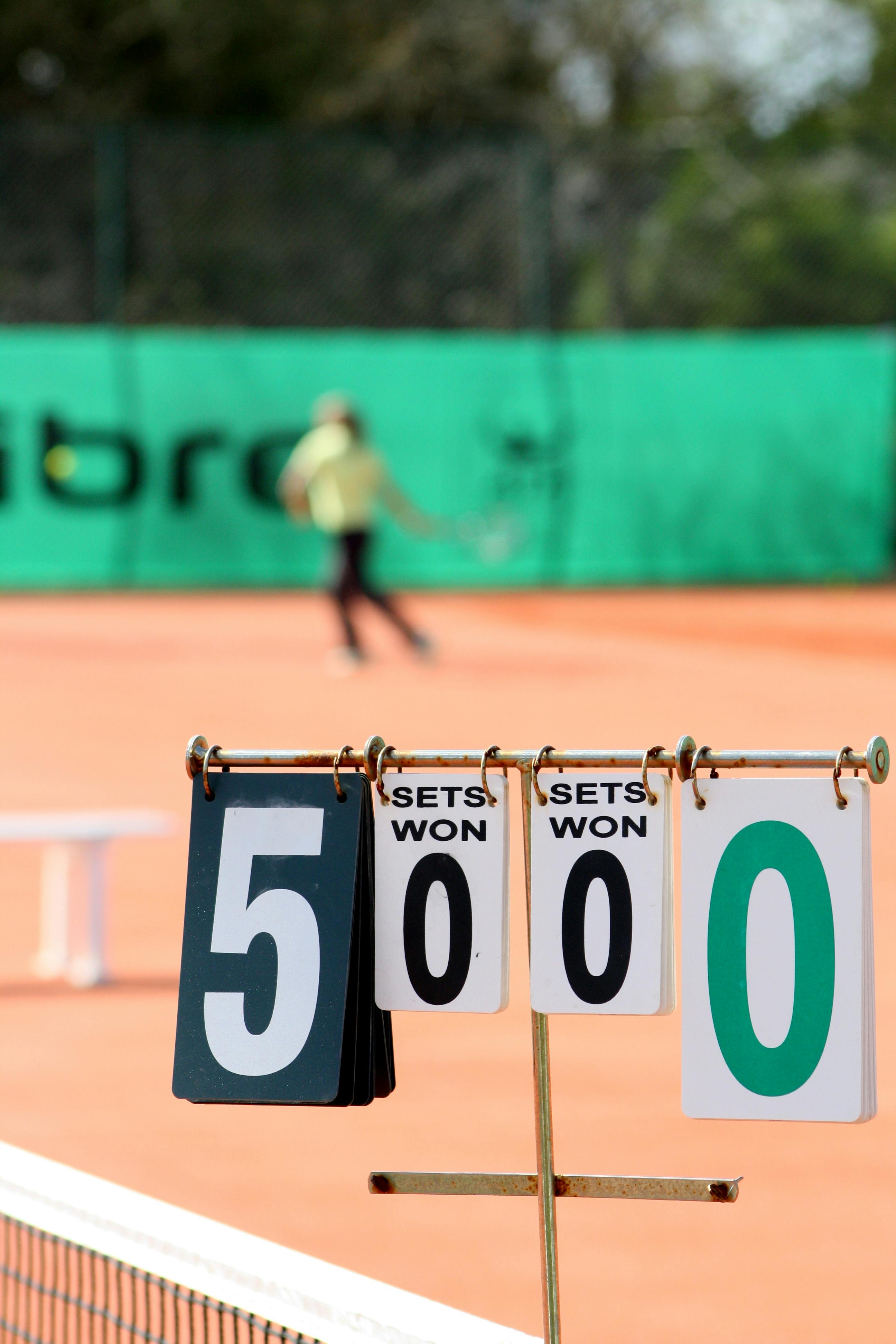 Close-up of tennis scoreboard with blurred player in action on clay court.