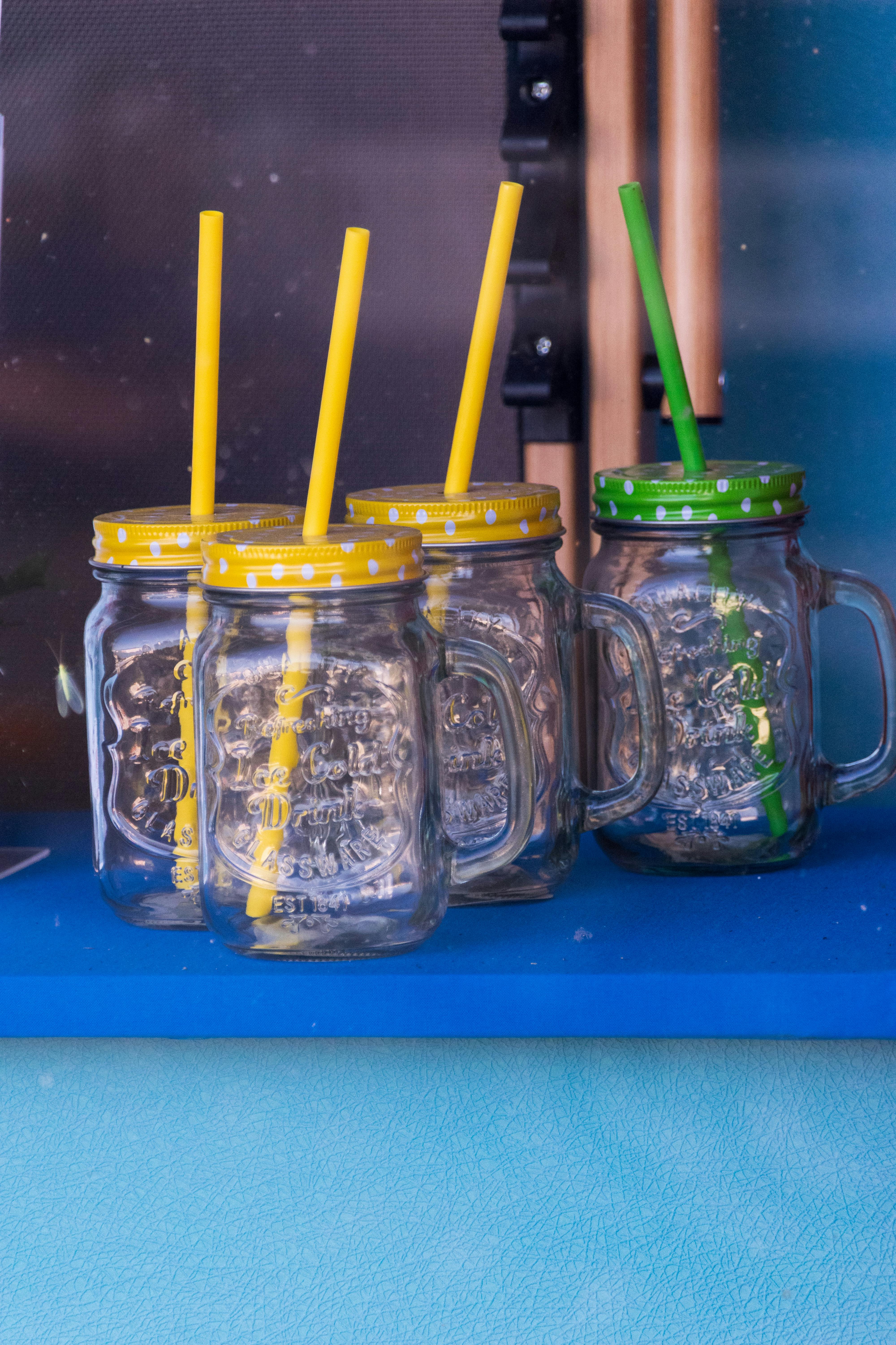 Four mason jar glasses with vibrant lids and straws on a blue shelf.