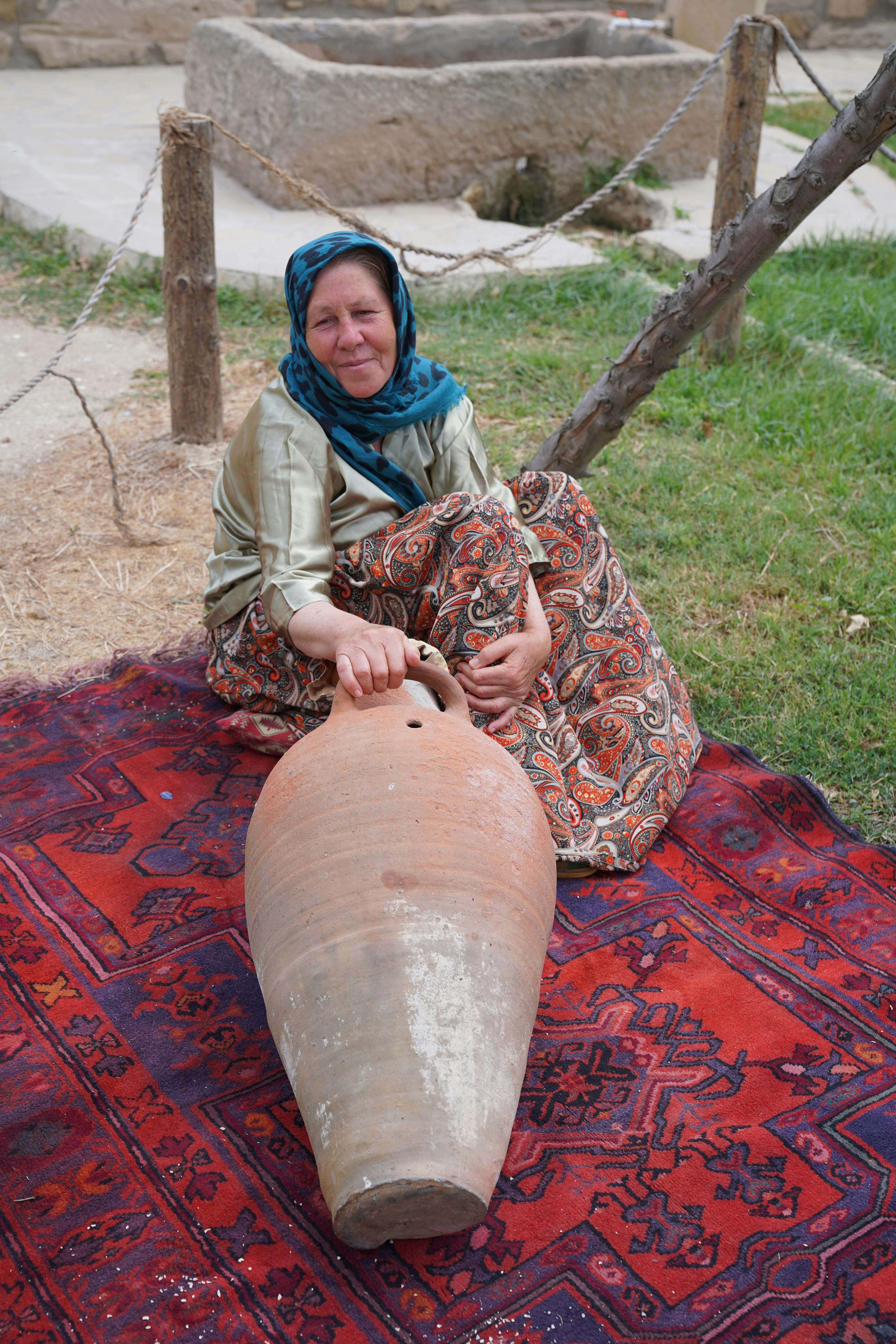 Woman sitting on a vibrant rug outdoors with a large clay vessel, showcasing cultural heritage.