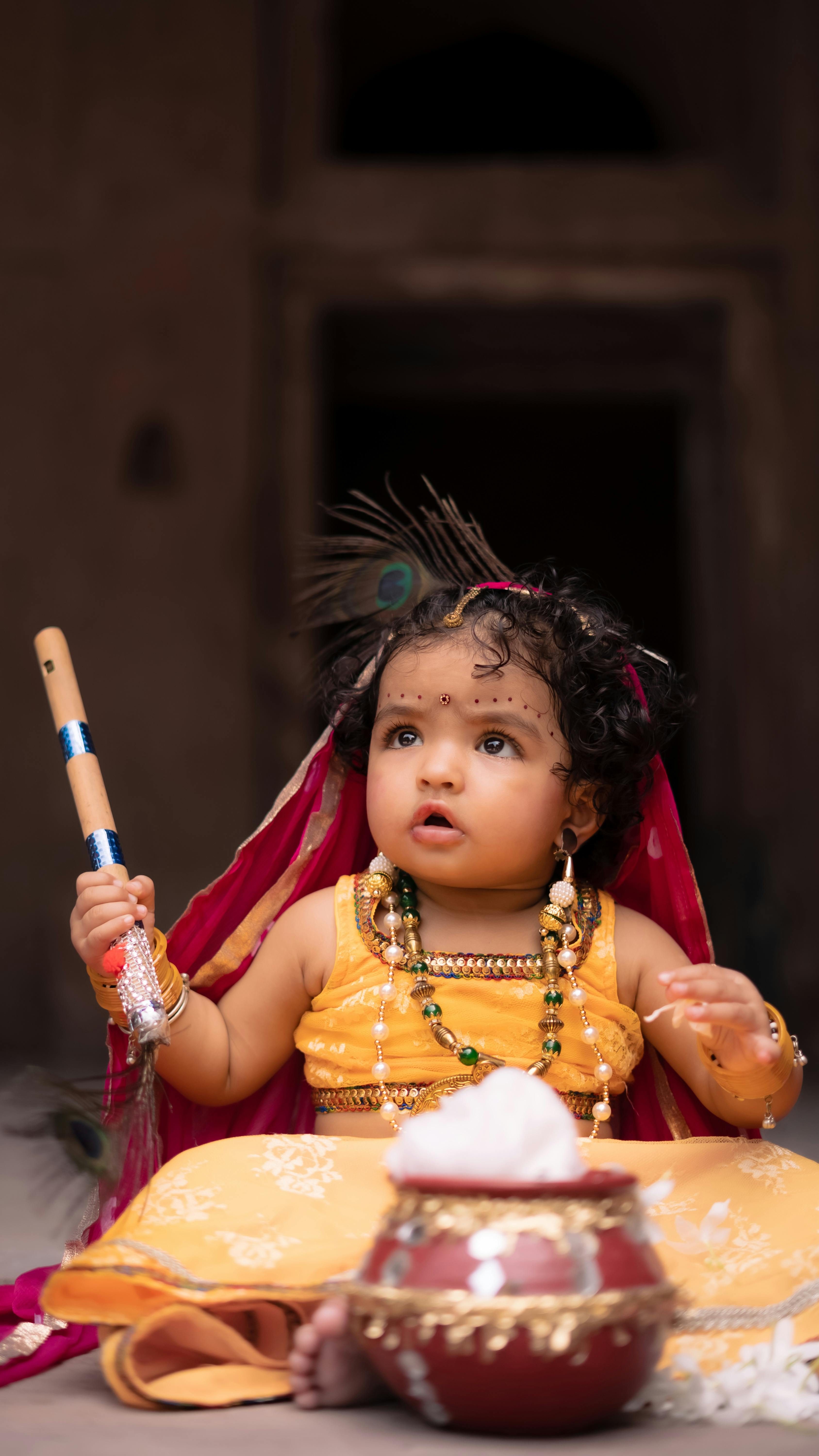 Cute baby dressed in vibrant traditional Indian attire, holding a flute with a clay pot nearby.